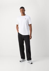 Hvid kortærmet t-shirt med et lille rødt logo, parret med sorte løstsiddende jeans og grå sportsko med sorte accenter.