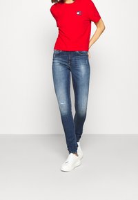 Kobieta w czerwonej koszulce Tommy Jeans, niebieskich wąskich dżinsach z delikatnymi przetarciami i białych trampkach, stojąca na jednolitym tle.