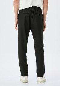 Zwarte stretch joggerbroeken met een elastische tailleband, twee achterzakken en taps toelopende pijpen, gecombineerd met witte sneakers op een neutrale achtergrond.