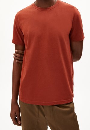 Mężczyzna ubrany w rdzawy t-shirt z krótkim rękawem i brązowe spodnie, z jednym ramieniem zgiętym i ręką chwytającą przeciwne ramię.