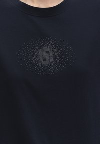 Gros plan d’une chemise bleu foncé avec un motif circulaire de petits points argentés formant les lettres « B » et « R » sur la poitrine.
