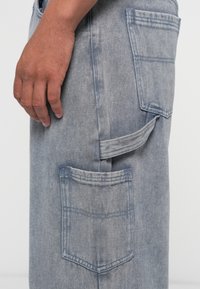 Lichtblauwe denim cargobroek met opgestikte zakken, zichtbare stiksels en een lusaccent aan de zijkant, die een gestructureerde stof toont.