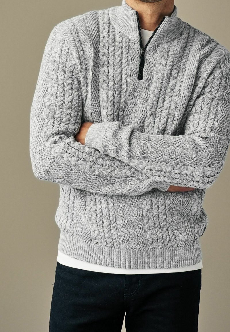 Grauer gestrickter Pullover mit halbem Reißverschluss-Kragen, der strukturierte Zopfmuster und gerippte Bündchen sowie Saum aufweist. Über einem weißen Hemd getragen.