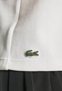 Tissu blanc texturé avec un petit logo crocodile vert près de l'ourlet, au-dessus d'un tissu noir plissé.