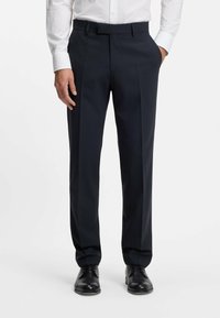 Pantaloni sartoriali blu in un tessuto liscio, con pieghe frontali e frontale piatto. Abbinati a scarpe di pelle nere.