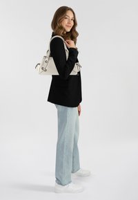 Borsa a tracolla triangolare bianca con hardware nero a contrasto; il modello indossa una giacca nera e jeans svasati azzurri.