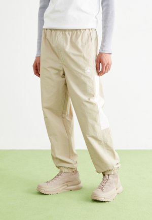 Persoon draagt beige joggingbroek met elastische taille, wit zijpaneel en beige hoge sneakers op een lichtgroene vloer.