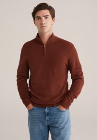 Rostfarbener Strickpullover mit einem Viertelreißverschluss, gerippten Bündchen und einem strukturierten Muster, kombiniert mit hellblauen Jeans.