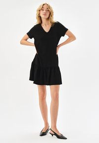 Czarna, prążkowana sukienka z dekoltem w serek, krótkimi rękawami i rozkloszowanym dołem. Zestawiona z eleganckimi czarnymi butami na obcasie typu slingback. Gładka faktura.
