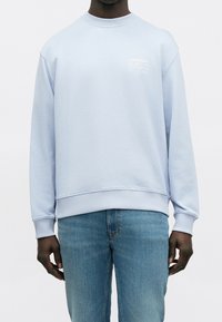 Homme portant un sweat Calvin Klein bleu clair avec poignets et ourlet côtelés, associé à un jean bleu moyen, debout sur un fond uni.