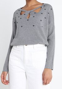 Vrouw in een grijze lange mouwen trui met uitsnijdingen en kleine zwarte lippenafdrukken, gecombineerd met hoog getailleerde witte broek met grote zakken.