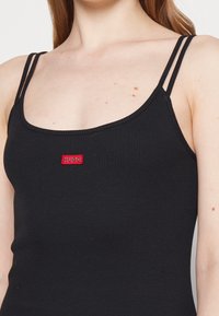 Mustat ribbed-hihattomat toppi ohutolkaimilla, jossa on punainen "HUGO"-logo-patch edessä. Materiaali on tiukka istuinen ja teksturoitu pinta.