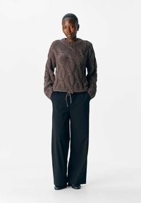 Maglione marrone a maglia con scollo rotondo e arricciature all'orlo, abbinato a pantaloni neri larghi e scarpe nere. Sfondo minimalista.
