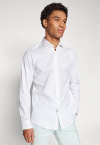 Camisa blanca de manga larga con botones, confeccionada en algodón y con un cuello clásico. Presenta un dobladillo redondeado y un ligero brillo. Accentos de botones cerca de los puños.