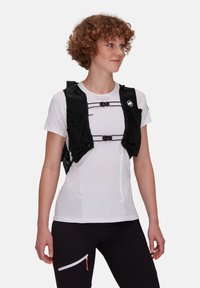 Czarna kamizelka biegowa z siateczkowymi akcentami, regulowanymi paskami i wieloma kieszeniami, noszona na dopasowanej białej koszulce i ciemnych spodniach z kieszenią na zamek.