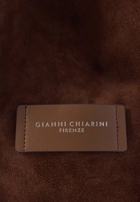 Hnedé semišové pozadie s obdĺžnikovou koženou štítkom prišitým, ktorý obsahuje vyrazený strieborný text: "GIANNI CHIARINI FIRENZE."