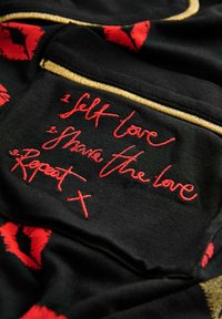 Must mustriba mustriba punased huulte mustrid; sisaldab punases tikandit tekstiga: "1. Armasta ennast, 2. Jagage armastust, 3. Korrake" kuldse äärisega.