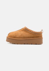 Zuecos de ante color tan sin cordones, con un acento textil estampado en el cuello, suela de goma elevada y un lazo en la parte posterior para facilitar el uso.