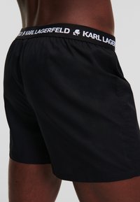 Crne kupaće gaće izrađene od laganog materijala, s elastičnom strukom i bijelim logotipom "KARL LAGERFELD". Minimalistički dizajn.