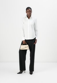 Weißes Hemd mit Knöpfen und einer Tasche, kombiniert mit schwarzen weiten Hosen und schwarzen High Heels. In der Hand eine cremefarbene Handtasche mit einem charakteristischen Detail.