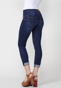 Jeans de mezclilla azul oscuro con un corte ajustado, puños enrollados y bolsillos traseros adornados. Combinados con sandalias de plataforma marrones con tacón.