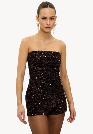 Femme portant une combinaison courte sans bretelles, ornée de sequins foncés, avec de grandes boucles d'oreilles en or, posant devant un fond blanc uni.