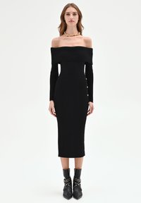 Schwarzes, geripptes Off-Shoulder-Kleid mit langen Ärmeln, figurbetontem Schnitt und seitlichen Knöpfen. Kombiniert mit schwarzen Stiefeletten mit Nietenakzenten.