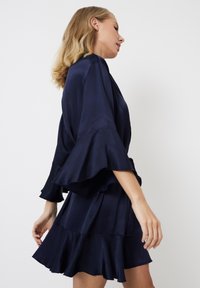 Robe en satin bleu marine avec des manches évasées, des volants superposés et un léger éclat. Présente une coupe décontractée et un design fluide pour un confort optimal.