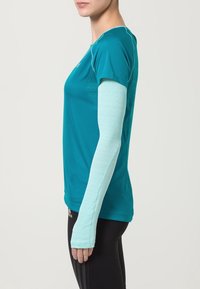 Turkoosi urheilupaita, jossa on lyhyet hihat ja pitkät, vaaleansiniset hihansuut. Teksturoidusta kankaasta valmistettu ja slim fit -suunnittelu, täydellinen aktiiviseen käyttöön.