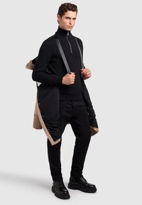 Suéter negro de cuello alto con cremallera, acompañado de un abrigo largo beige con detalles negros. Pantalones negros de corte slim y botas negras gruesas.