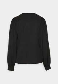 Blouse noire à manches longues avec épaules bouffantes et poignets boutonnés, vue de dos sur un fond gris clair uni.