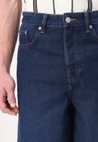 Jean en denim de couleur bleu foncé, avec une coupe classique, des jambes droites, des coutures contrastées et des poches avant avec un bouton en métal argenté.