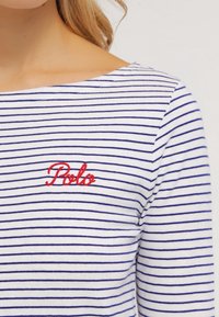 Randig långärmad topp i vitt och marinblått. Har en rund halsringning och broderad röd "Polo"-logotyp på bröstet. Mjuk bomullstextur.