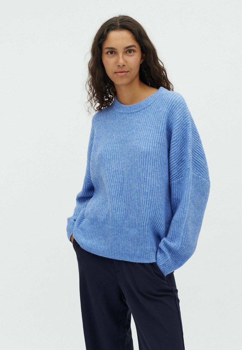 Sweater côtelé bleu clair avec des épaules tombantes et un col rond, fabriqué en tissu doux et texturé, associé à un pantalon noir.