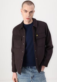 Mørkebrun knap-op jakke med to brystlommer, sølvknapper og et lille gult logo. Sæt sammen med en marineblå sweater og blå jeans.