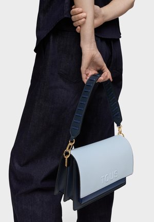 Persona con ropa oscura sosteniendo un bolso TOUS azul claro y azul marino con una correa ancha y texturizada y herrajes dorados a su lado.