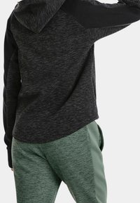 Sudadera con capucha en tejido jaspeado gris oscuro, mangas negras, combinada con pantalones de chándal verde claro. Textura suave, corte casual y capucha ajustable.