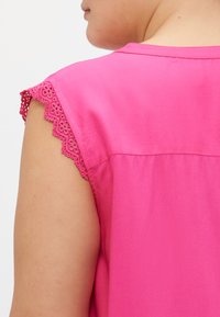 Blusa rosa con maniche corte, caratterizzata da un bordo in pizzo scallop. Il tessuto sembra morbido e leggero, con un design del colletto semplice.
