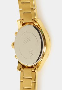 Montre en métal doré avec un bracelet à maillons, boîtier rond et dos poli argenté. Elle possède deux boutons latéraux et une couronne texturée.