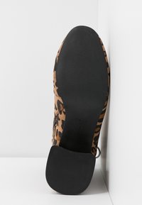 Semelle d'une botte cheville à imprimé léopard avec un talon plat noir, positionnée debout contre un mur blanc dans un coin.