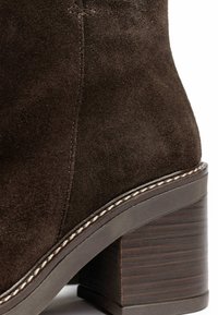 Brown suede ankle boot featuring a textured heel, detailed stitching along the sole, and a chunky block heel design.