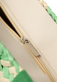 Baumwolltasche mit einem cremefarbenen Stoffkörper, grünen geflochtenen Akzenten und einem hellbeigen Reißverschluss mit einem silbernen Ziehgriff.