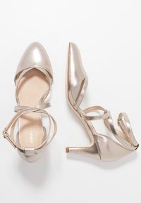 Metalické zlaté boty na vysokém podpatku s špičatou špičkou, překříženými pásky kolem kotníku a hladkým povrchem. Výška podpatku je přibližně 5 cm.
