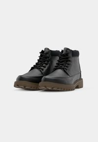Botas de cuero negro hasta el tobillo con suelas robustas, cuello acolchado y suelas de goma marrón contrastante. Presentan un diseño de cordones y detalles de costura.