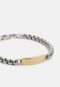 Pulseira de corrente prateada com uma placa retangular dourada em relevo com a inscrição "GUESS." Textura suave, design interligado e fecho seguro.