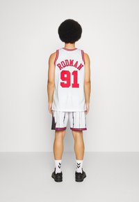 Tricou de baschet din plasă albă cu accente roșii, text "RODMAN" și numărul "91." Asortat cu pantaloni scurți cu model zigzag. Adidași negri.
