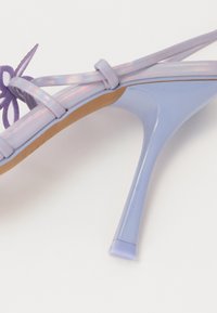 Sandale à talons hauts avec lanières en violet clair irisé, silhouette élégante, bout pointu et un accent délicat de nœud sur la lanière de la cheville.