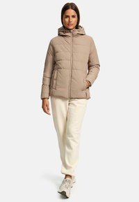 Beige gewatteerde jas met een capuchon, ritssluiting en voorzakken, uitgerust met een gewatteerd ontwerp en elastische manchetten. Gepaarde met crèmekleurige joggingbroek.