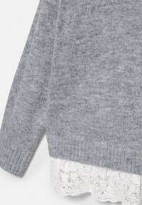 Manche et ourlet de pull gris en maille superposés à un tissu en dentelle blanche à motif floral en dessous.