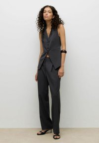 Dopasowany szary zestaw kamizelki i spodni z głębokim dekoltem w serek, pięcioma guzikami i kieszeniami po bokach, w połączeniu z czarnymi sandałami na obcasie.